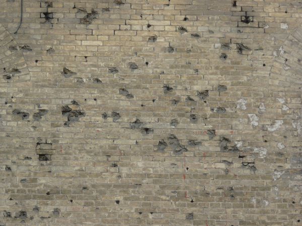 Brick wall in light brown and grey tones with chipped, damaged surface.