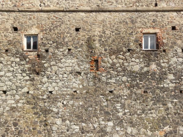 Medieval stone wall with rough texture and windows and holes in surface.