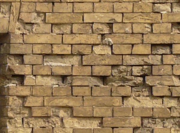 Wall texture made up of yellow bricks in various degrees of releif and stages of erosion. Several tall, narrow windows are visible in the brick, and large grey stones line the bottom.