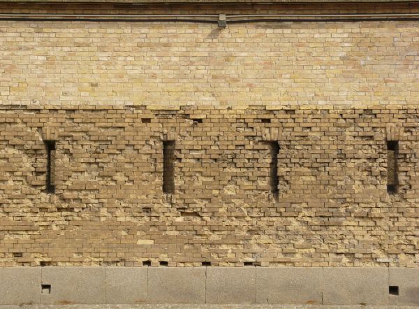 Wall texture made up of yellow bricks in various degrees of releif and stages of erosion. Several tall, narrow windows are visible in the brick, and large grey stones line the bottom.