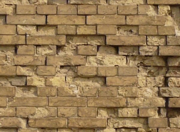 Wall texture made up of yellow bricks in various degrees of releif and stages of erosion. Several tall, narrow windows are visible in the brick, and large grey stones line the bottom.