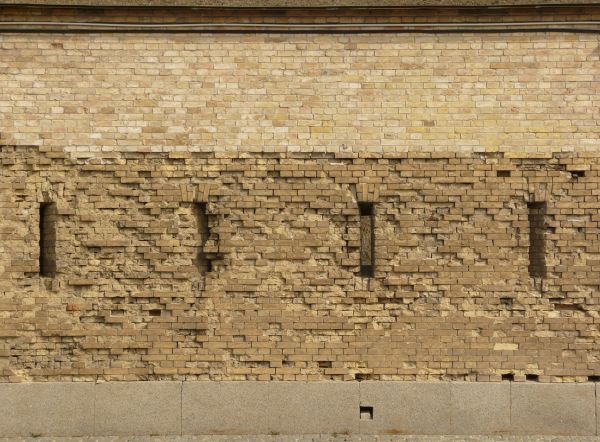 Wall texture made up of yellow bricks in various degrees of releif and stages of erosion. Several tall, narrow windows are visible in the brick, and large grey stones line the bottom.