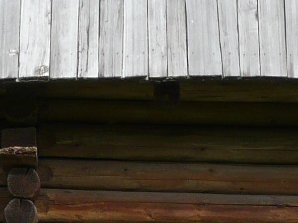 Small, old cabin with stone base and walls of wooden logs. Roof is made of grey, wooden planks.