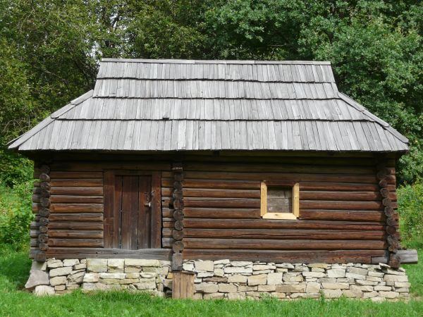 Small, old cabin with stone base and walls of wooden logs. Roof is made of grey, wooden planks.