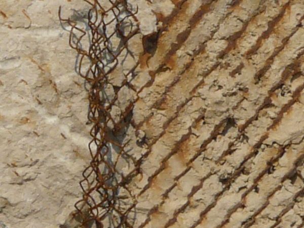 Very rough, damaged concrete in light beige color settled over rusty, metal mesh.