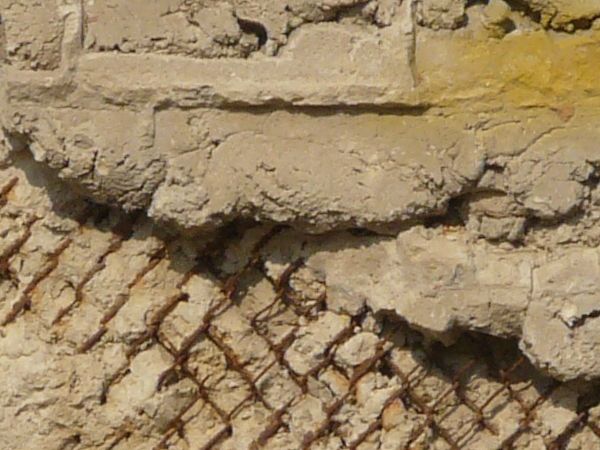 Very rough, damaged concrete in light beige color settled over rusty, metal mesh.