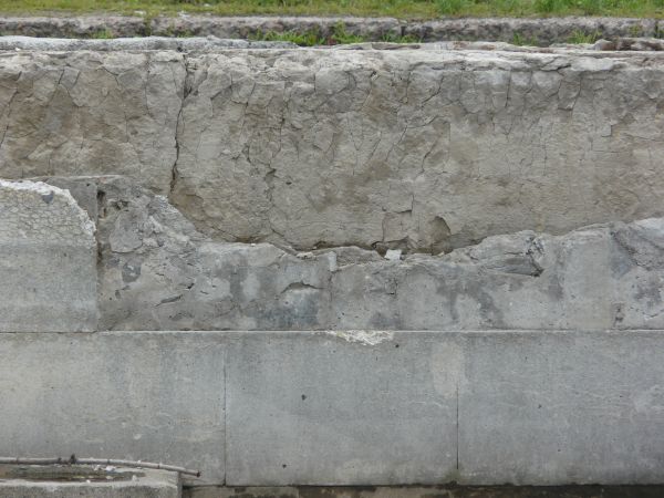 Short wall of grey concrete with large cracks and very damaged, worn surface.