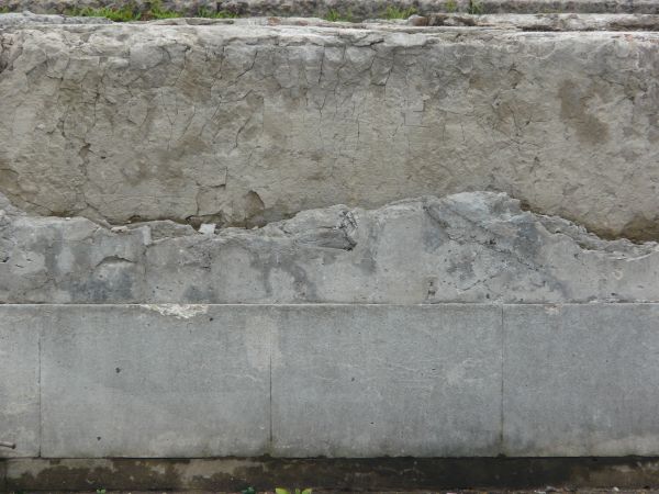 Short wall of grey concrete with large cracks and very damaged, worn surface.