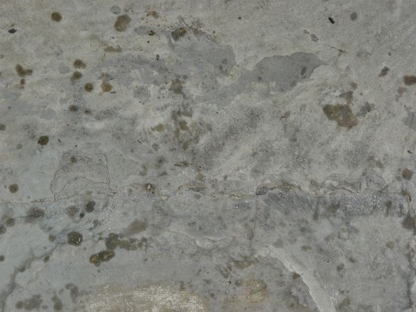 Flat concrete texture in grey tones with random, dark spots across surface.
