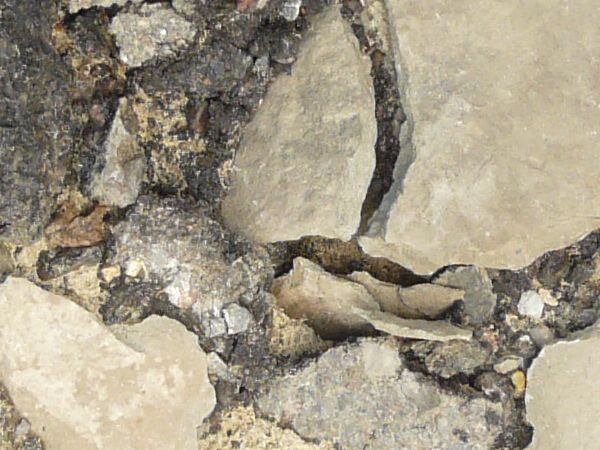 Road texture, broken completely into large and small chips of asphalt, with light, sandy earth and dark rock visible underneath.