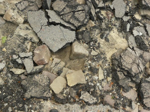 Road texture, broken completely into large and small chips of asphalt, with light, sandy earth and dark rock visible underneath.