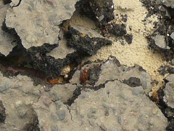 Road texture, broken completely into large and small chips of asphalt, with light, sandy earth and dark rock visible underneath.