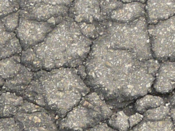 Road texture, rough and grey in color, with various thin cracks and holes worn into most of the surface. A small area of dark, defined cracks is also visible.