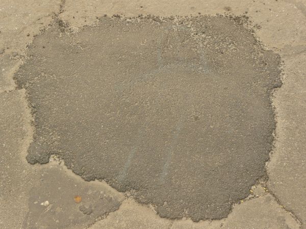 Road texture, sandy brown in color, with a large, irregularly shaped patch of dark asphalt on the center.