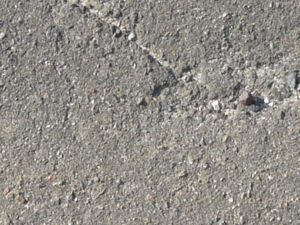 Road texture, grey in color, with long, defined cracks, some crumbling areas, and spots of fine dust and small rocks.