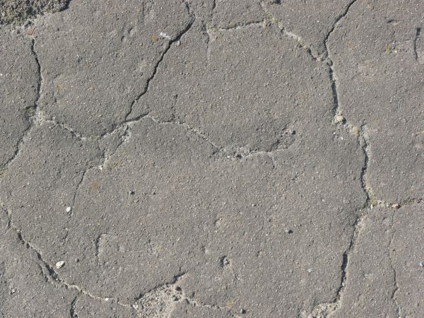 Road texture, grey in color, with long, defined cracks, some crumbling areas, and spots of fine dust and small rocks.