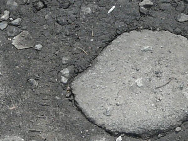 Road texture with a large hole worn into the surface, filled with dark, damp dirt and light-colored stones of various sizes scattered about.