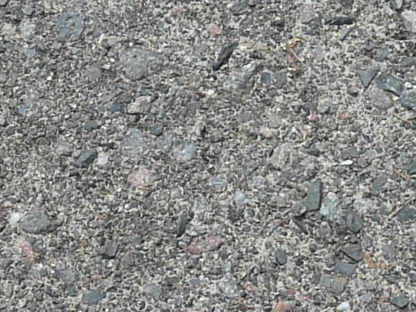 Road texture with a large hole cracked into the surface, which is filled with a coarse layer of small dark rocks. Extensive cracking and bits of weeds are present around the hole.