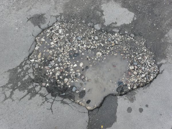 Road texture with a large, irregular hole cracked into the center that is filled with light and dark rocks, and a small amount of cloudy water. Much of the surface around the hole is cracked and damp.