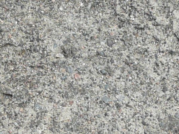 Road texture, dark grey in color, covered with cracks and a fine layer of light dust and small rocks. A circular, dust filled recession is in the center, and small bits of green weeds are visible.