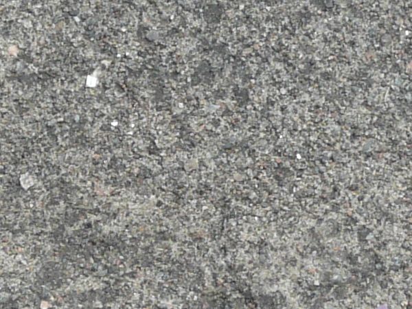 Road texture, dark grey in color, covered with cracks and a fine layer of light dust and small rocks. A circular, dust filled recession is in the center, and small bits of green weeds are visible.