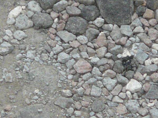 Road texture with large cracks and a irregular recession that is filled with various sizes of colored rocks. A manhole cover with a wavy pattern is nearby.