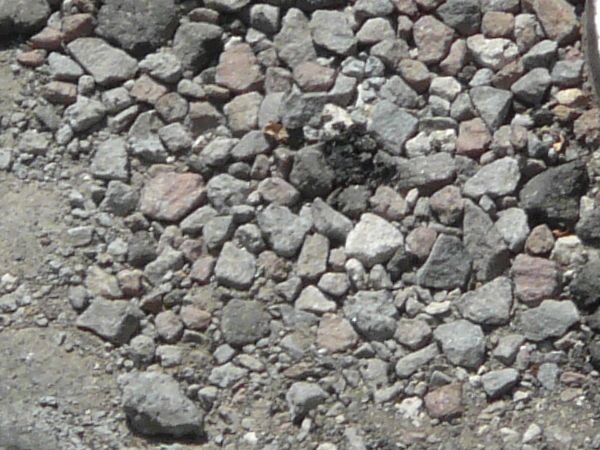 Road texture with large cracks and a irregular recession that is filled with various sizes of colored rocks. A manhole cover with a wavy pattern is nearby.