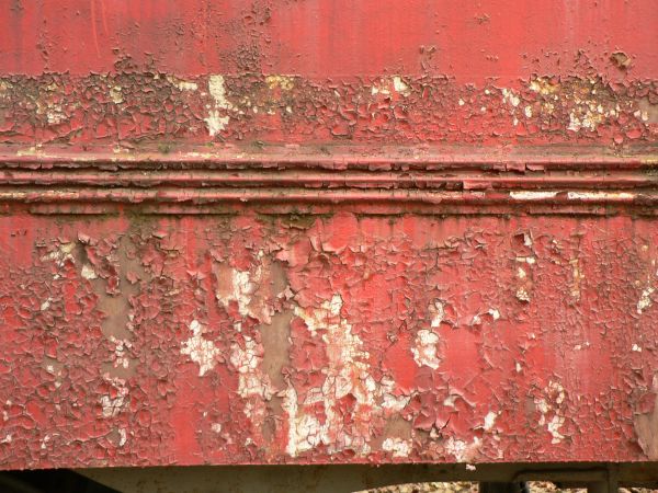 Red painted metal texture, with large chipping and peeling areas. Dark rusted areas are visible underneath the paint.