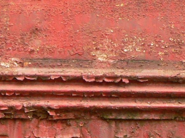 Red painted metal texture, with large chipping and peeling areas. Dark rusted areas are visible underneath the paint.