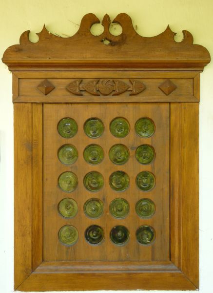 Green window texture, made up of small, separated circular panes, set in a large frame of smooth wood. Various patterns and tracery run along the top of the frame.