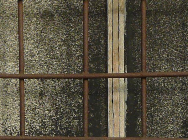 Brown window texture with a grid of thin bars set over it and a dark surface visible behind it. The window is surrounded by a wall of eroding tan brick.
