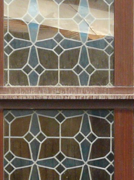Stained glass window texture with an arched top, bright and dark blue patterns in the glass and dark brown wood trim. The wall is of aging dark grey stone.