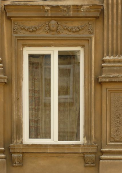 White window texture with a face and leaves molded above it, and various column and ledges surrounding it. A leaf patterned curtain is visible behind the glass