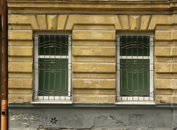 White windows texture with ornate black bars set over it, placed in a wall of smooth yellow stone. A surface of decaying grey stone runs below the windows.