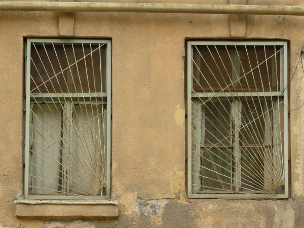 Light green windows texture, both covered by a sunburst pattern of bars, and set in a rough tan wall. Green and brown boards are visible behind the windows.