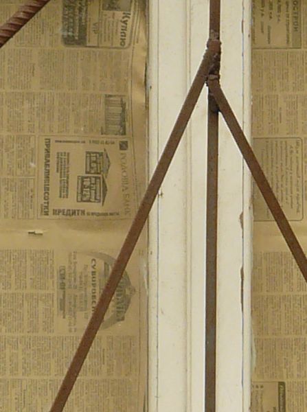 White window texture in a yellow wall, with an angular pattern of brown bars set over it. Yellowed newspaper is visible behind the bottom panes, and a strip of grey cement surrounds the window.