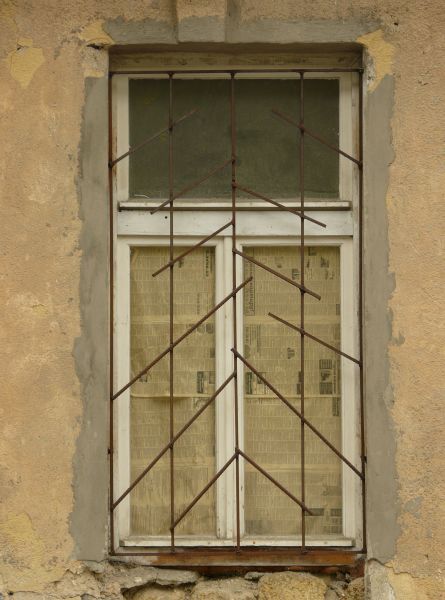 White window texture in a yellow wall, with an angular pattern of brown bars set over it. Yellowed newspaper is visible behind the bottom panes, and a strip of grey cement surrounds the window.