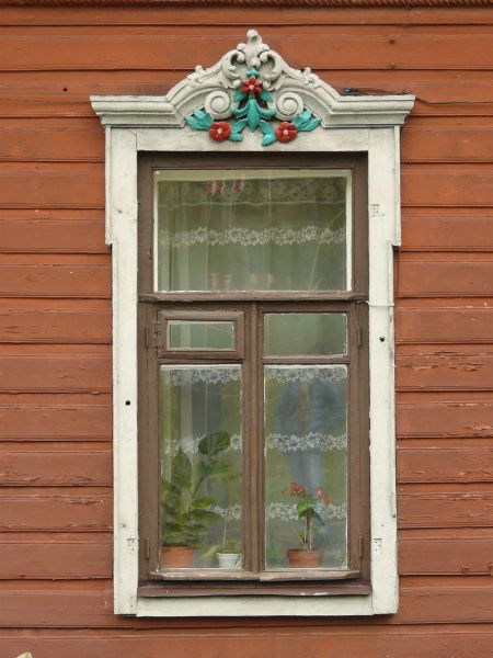 White window texture with brown trim and teal and red painted details at the top. A sheer white curtain and several potted plants are visible behind the glass.