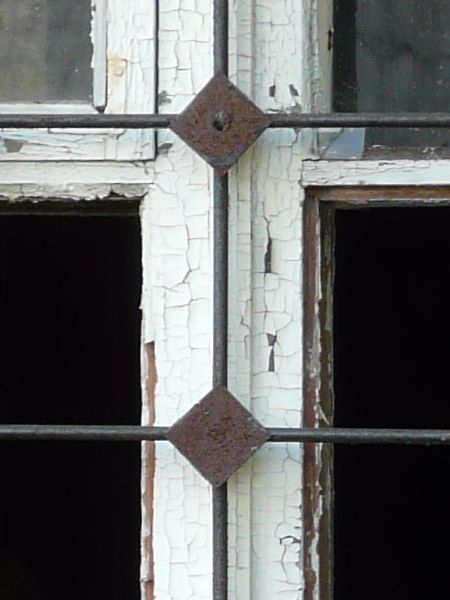 White window texture with chipping paint and missing panes, with a grid of iron bars set over it. The window is set in a brick wall painted red.