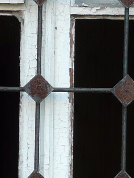 White window texture with chipping paint and missing panes, with a grid of iron bars set over it. The window is set in a brick wall painted red.