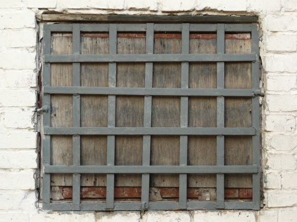 Window texture, boarded up and with a gird of wide grey bars set over it. The window is recessed in a brick wall painted white.