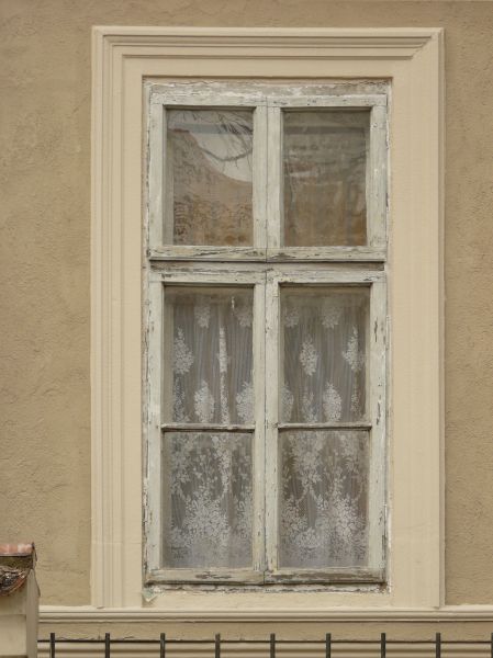 White window texture, with chipping paint and lace curtains visible behind the glass. The window is set in a rough beige wall.