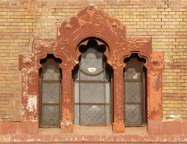 Red windows with tracery texture, divided into three sections in a wall of yellow and red brick. A fine grate is placed over some of the panes, and white material is visible behind them.