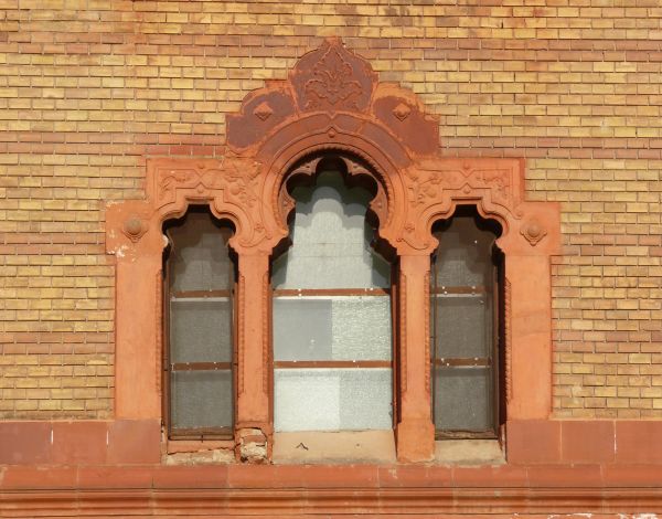 Red windows with tracery texture, divided into three sections in a wall of yellow and red brick. A fine grate is placed over some of the panes, and white material is visible behind them.