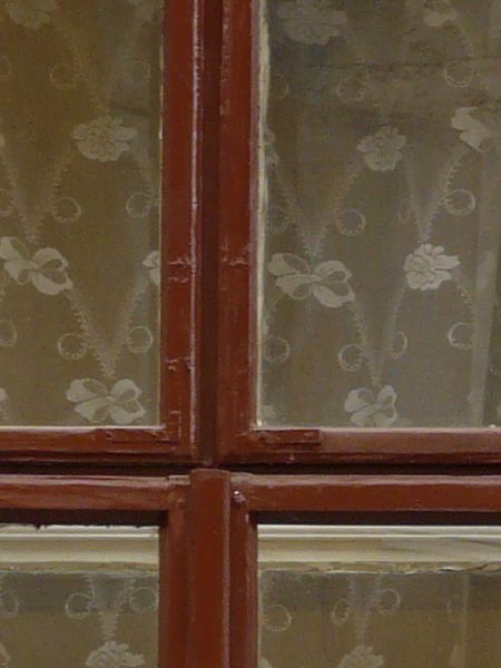 Red window texture, surrounded by an ornate frame with molding and ledges, and set in a reddish tan wall. A white lace curtain is visible behind the window.