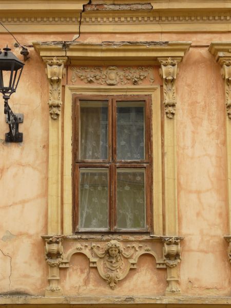 Red window texture, surrounded by an ornate frame with molding and ledges, and set in a reddish tan wall. A white lace curtain is visible behind the window.