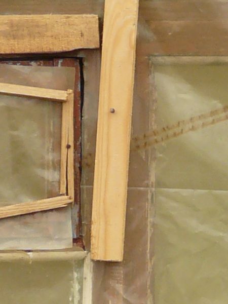 Plastic covered window texture, with scraps of brown and blue painted wood within the frame. The window is recessed in a red brick wall.