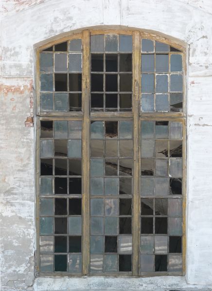 Arched window texture with yellow wood separating the panes, several of which are missing. A whitewashed wall surrounds the window, and large pieces of wood are visible behind it.