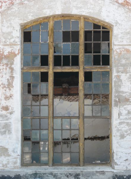 Arched window texture with yellow wood separating the panes, several of which are missing. A whitewashed wall surrounds the window, and large pieces of wood are visible behind it.