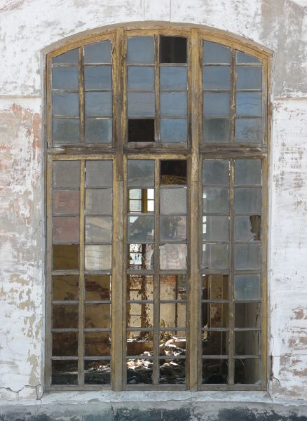 Arched window texture with aged yellow wood separating the small panes, several of which are missing. A whitewashed wall surrounds the window, and a room with debris is visible behind it.